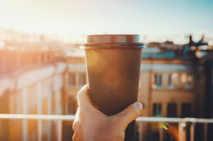 hands holding hot craft cup of coffee or tea in morning sunlight with view to blurred city background. Enjoy, lifestyle, take away breakfast concept. woman on the roof with hot drink